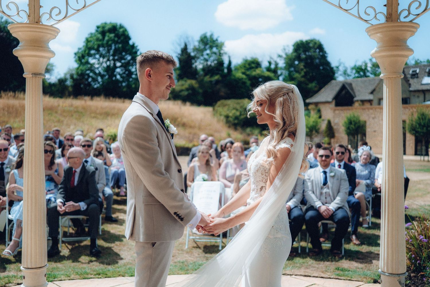 Summer wedding ceremony taking place under an ornate white gazebo.
