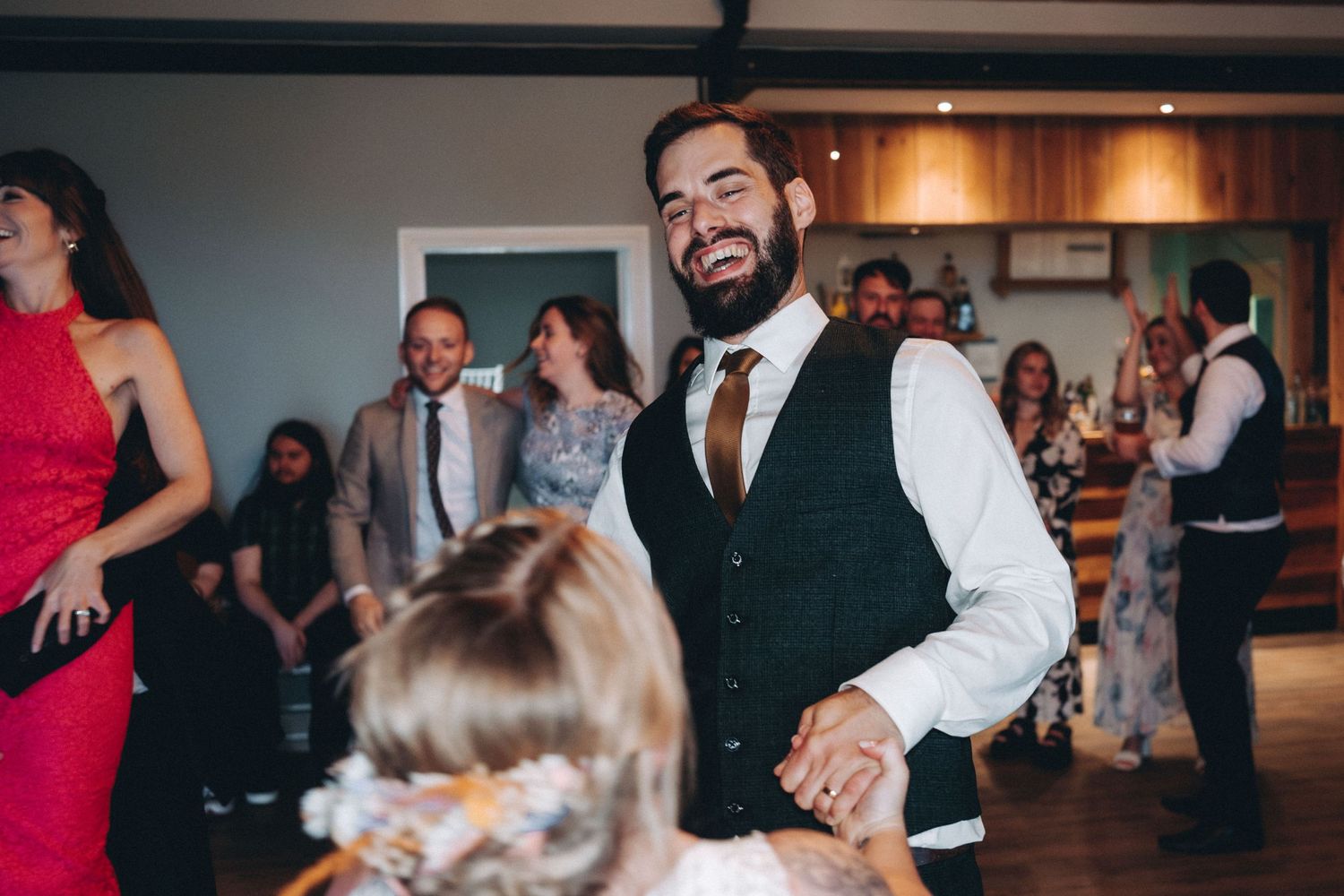 People dancing and celebrating at a wedding reception with guests in formal attire including a black vest and white shirt.