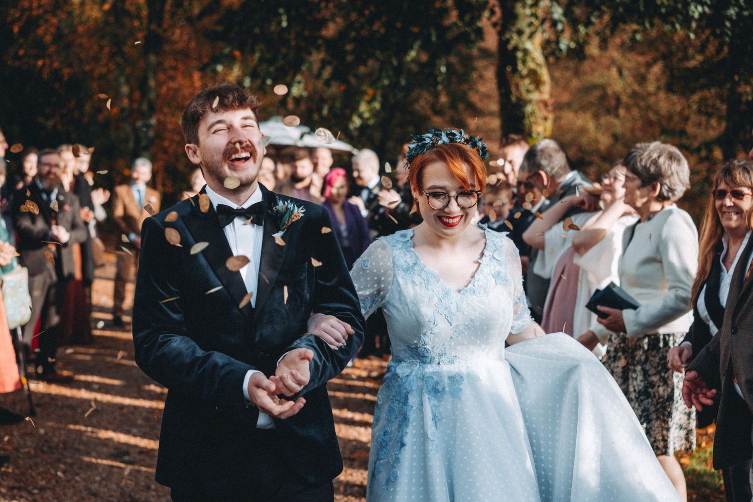 Wedding guests gather outdoors to celebrate as a couple walks down a leaf-covered path.