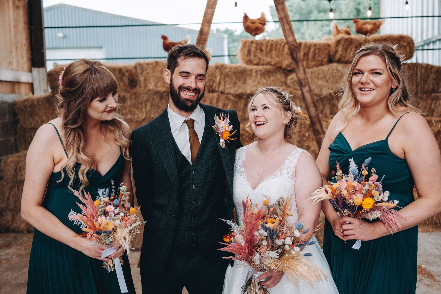 Wedding party in dark green dresses and a black suit holding vibrant fall-colored bouquets pose together at outdoor venue. Chickens.