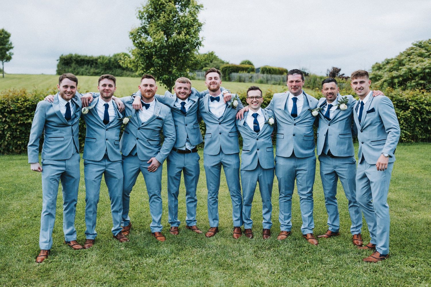 A group of groomsmen in matching light blue suits stand together on grass at an outdoor wedding venue.