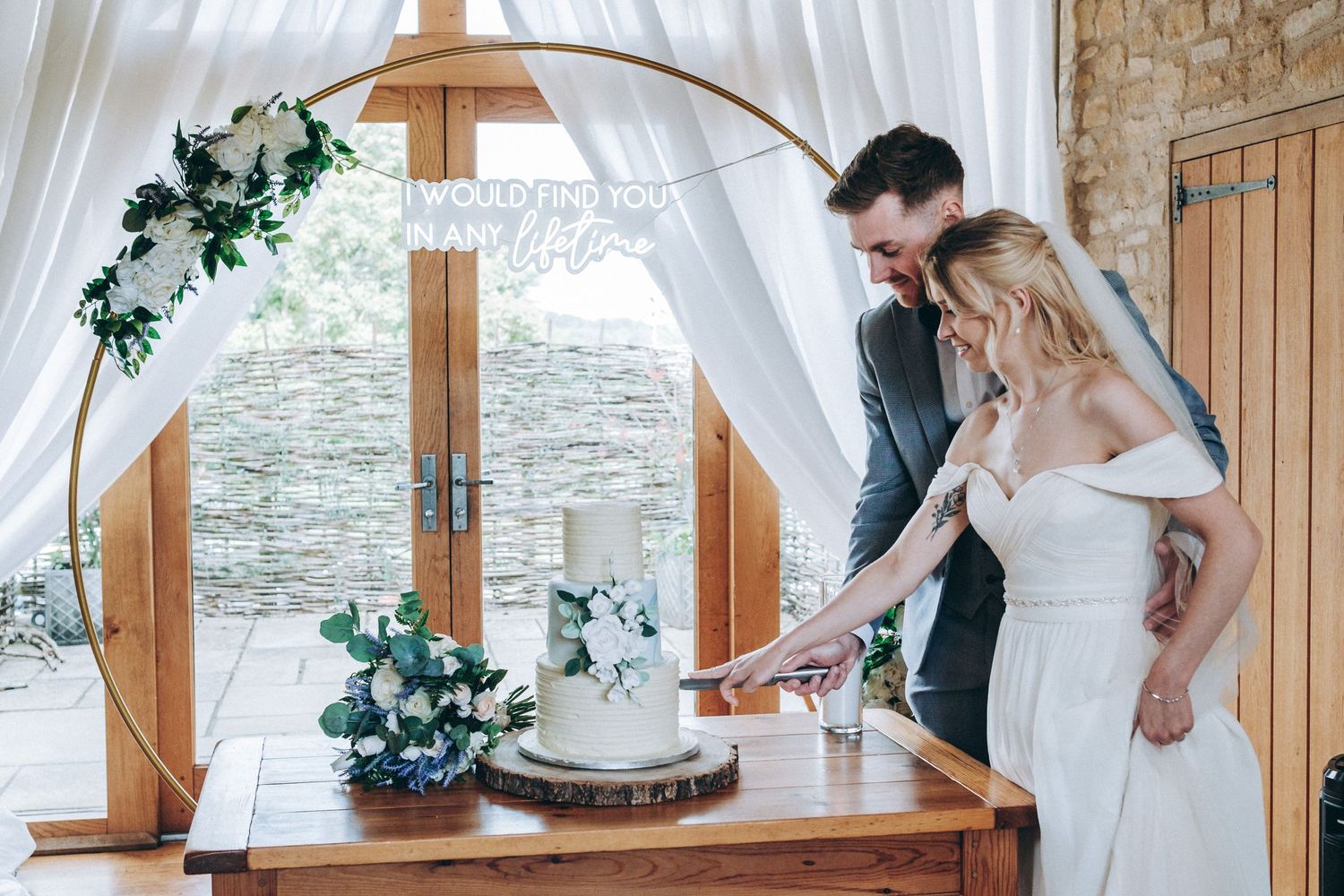 A romantic scene with a decorated archway and a white wedding cake being cut on a wooden table by the window.