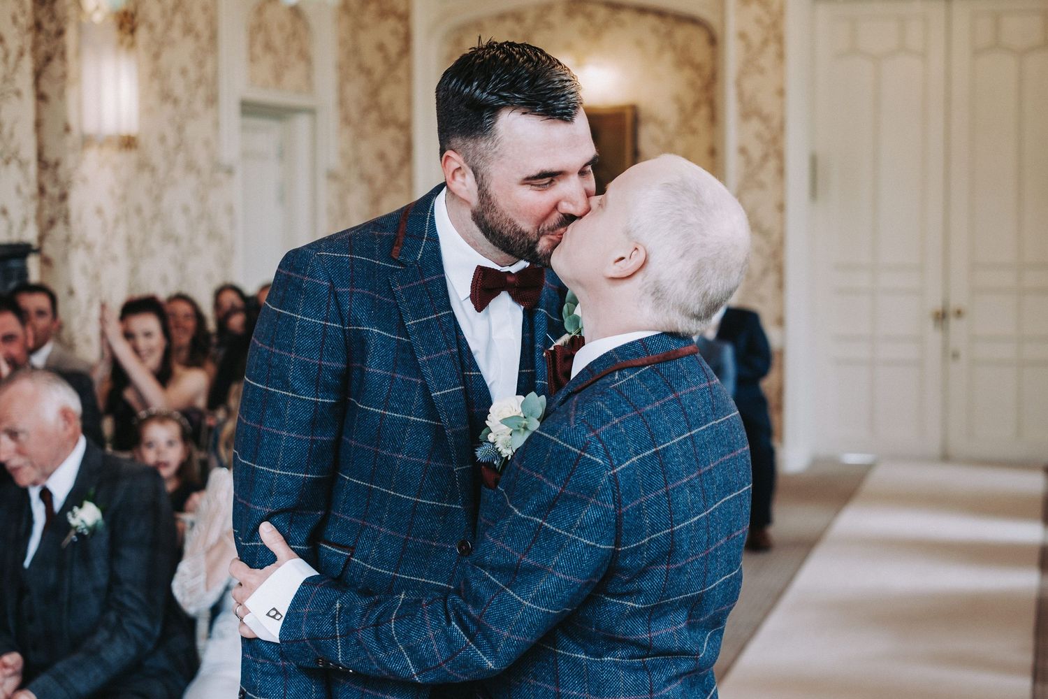 A couple in formal wedding attire share a tender kiss in an elegant venue with ornate wallpaper and decor.