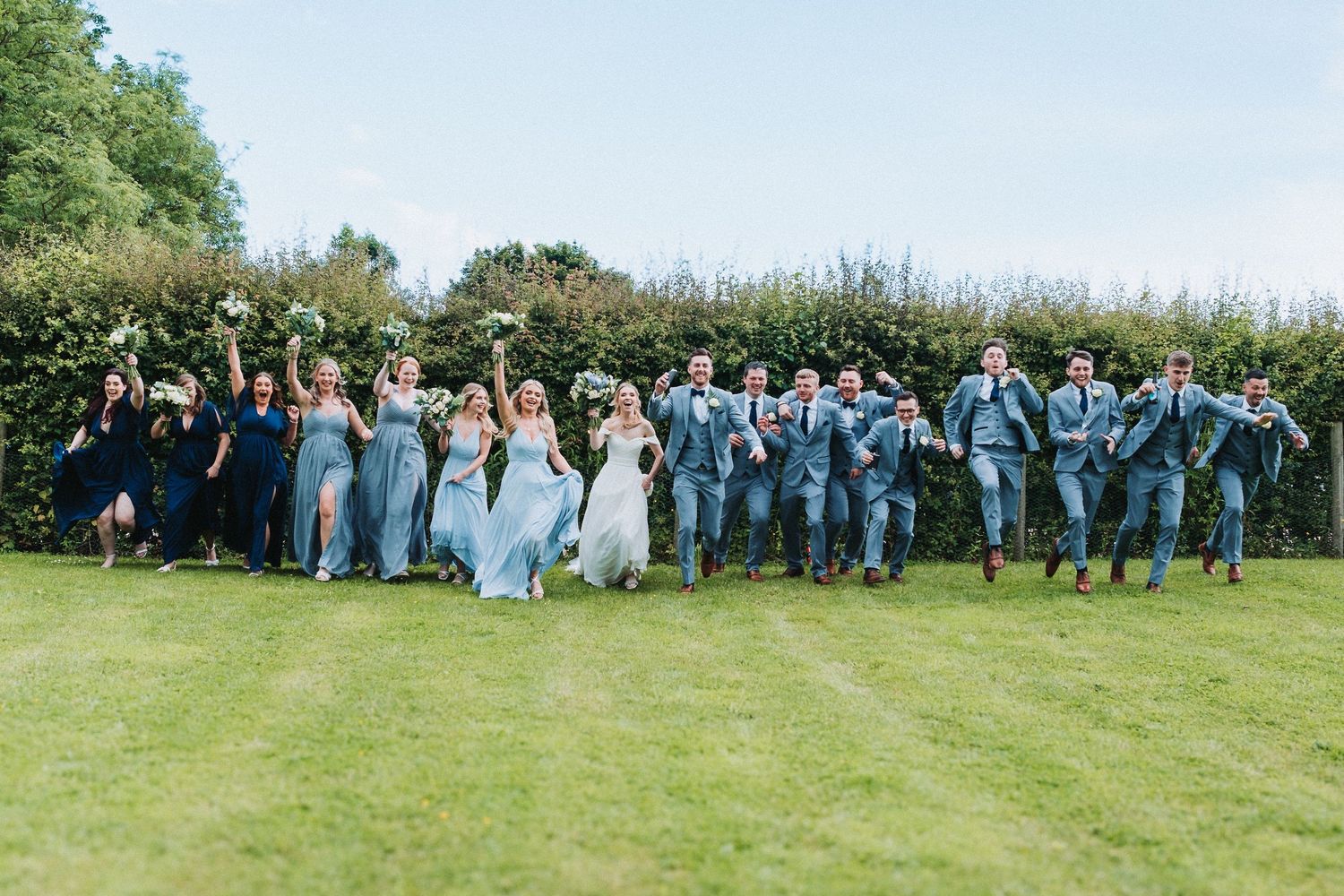 A large wedding party in blue and white attire jumps joyfully together on a green lawn during an outdoor celebration.