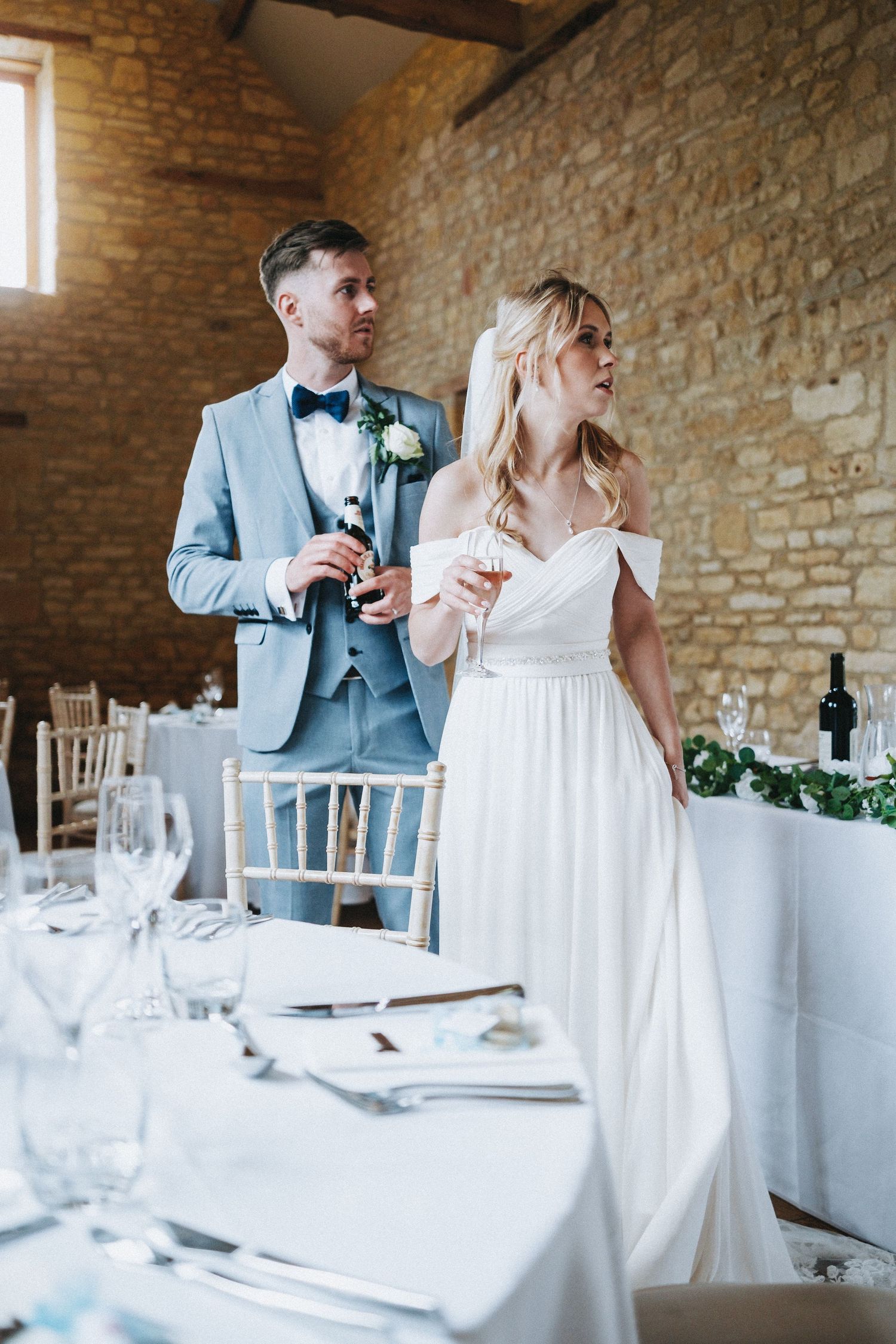 A couple stands together at an elegant wedding reception with stone walls and white table settings in a rustic venue.