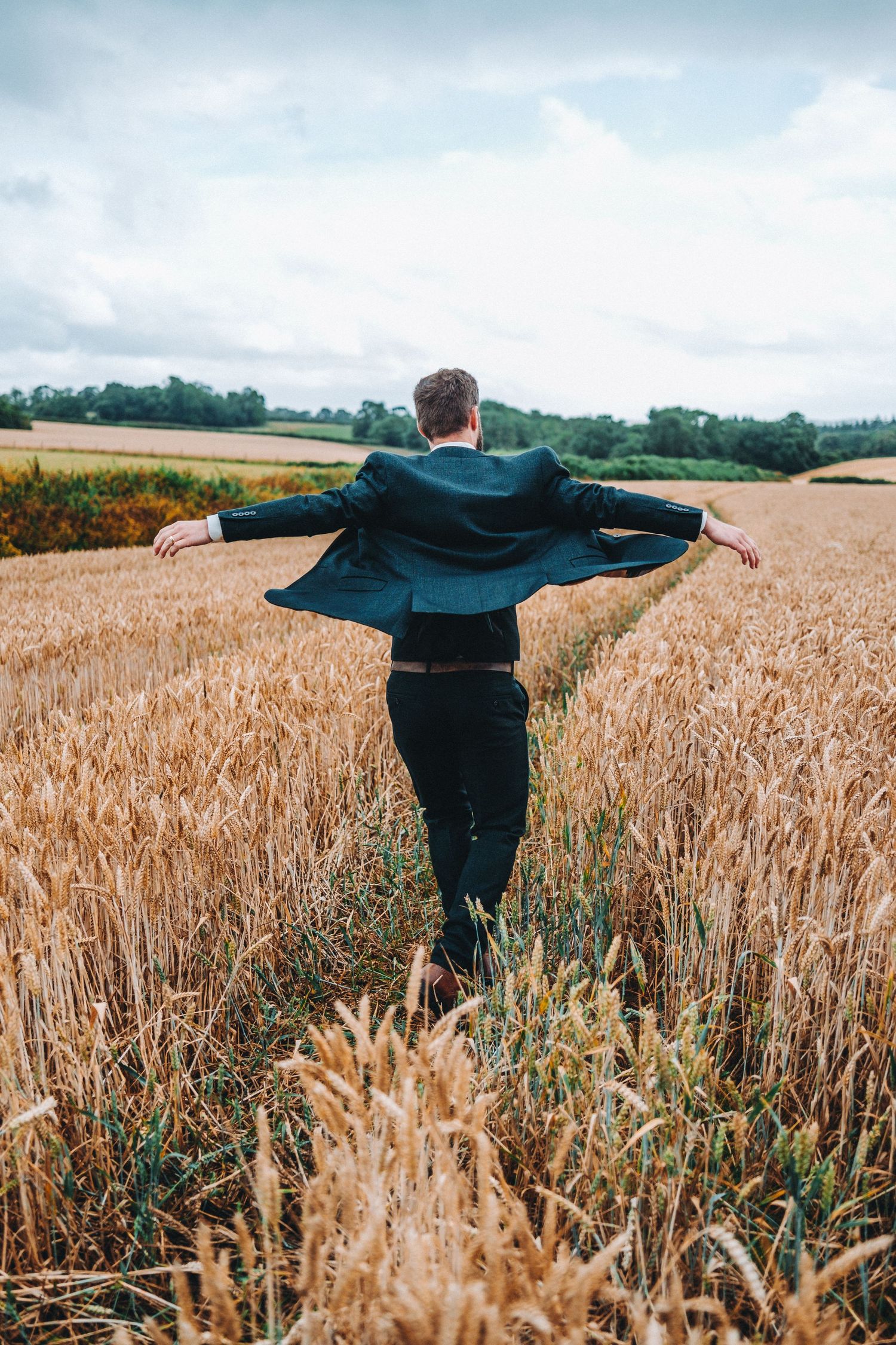 groom stands with outstretched arms in golden wheat field under cloudy sky.