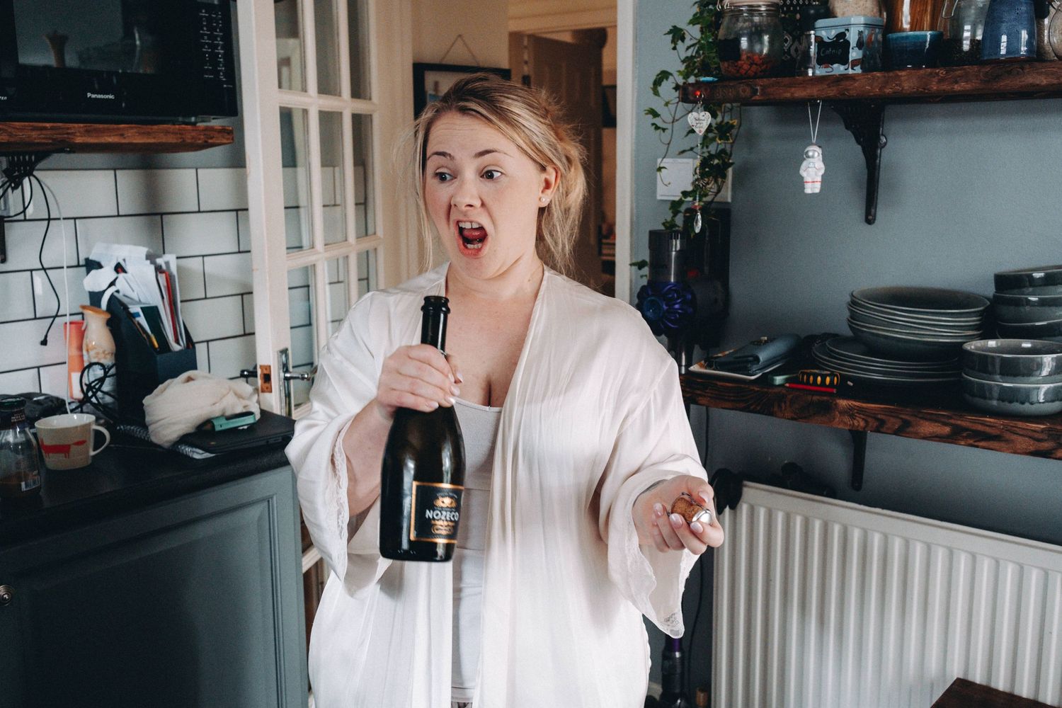 Bride in white robe making excited expression while holding wine bottle of champagne.