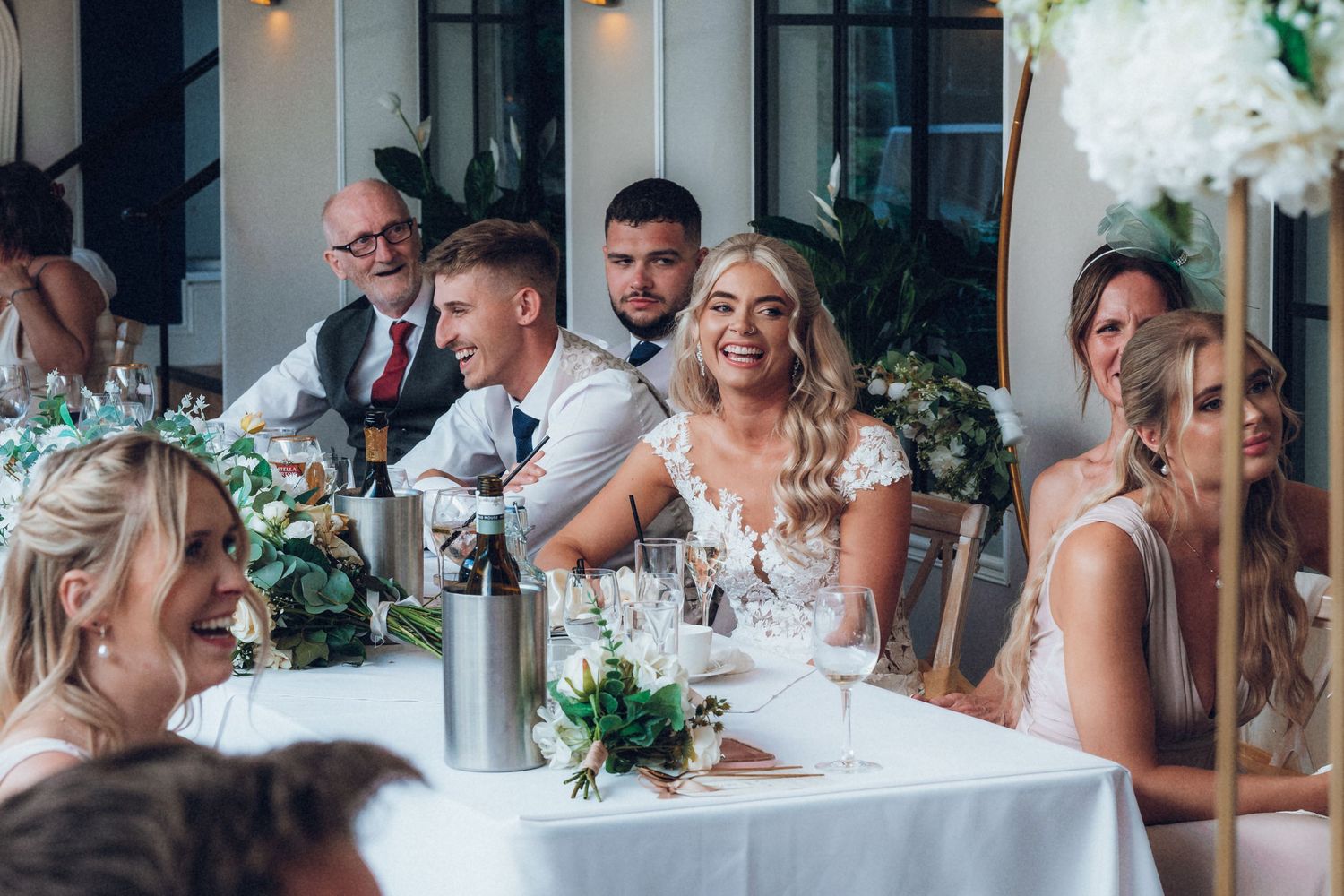 Wedding party seated at elegant white-clothed tables with floral centerpieces during reception.