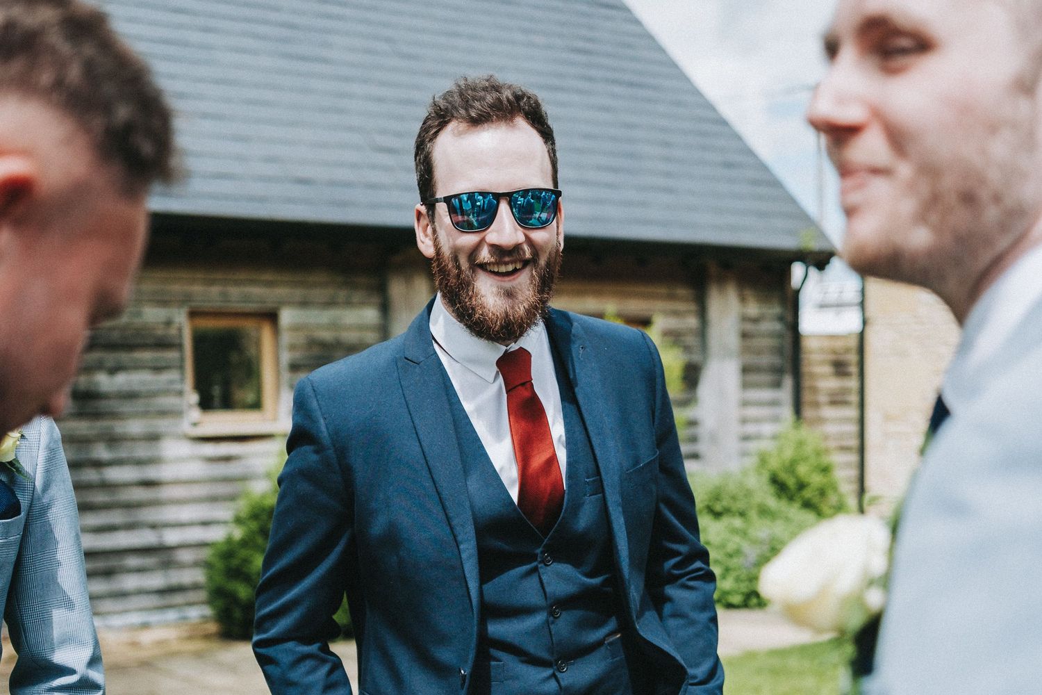 A guest in a navy blue suit with red tie smiles while socializing outdoors at a formal event.