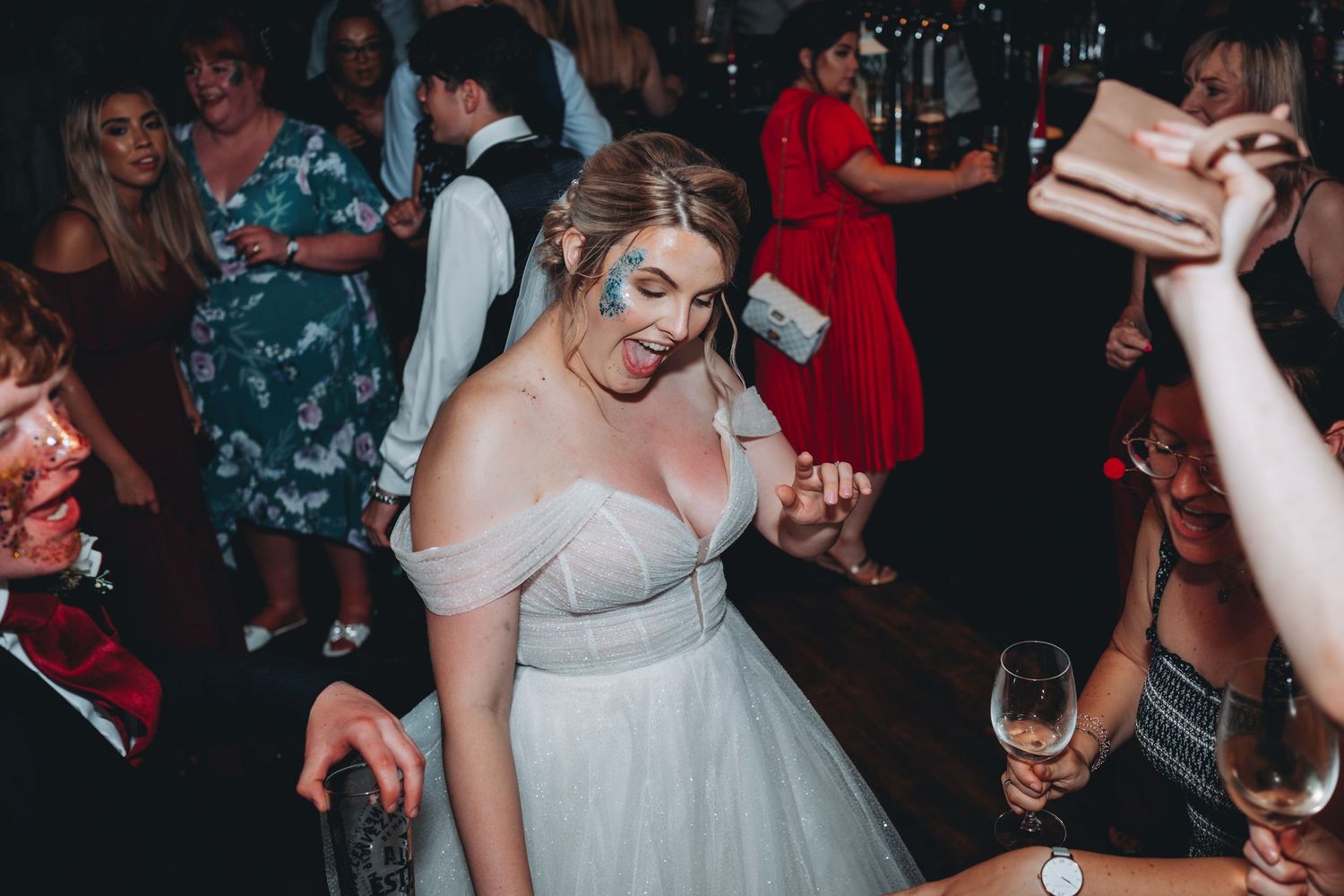 Bride celebrating and dancing at a party wearing an elegant off-shoulder white gown surrounded by other guests.