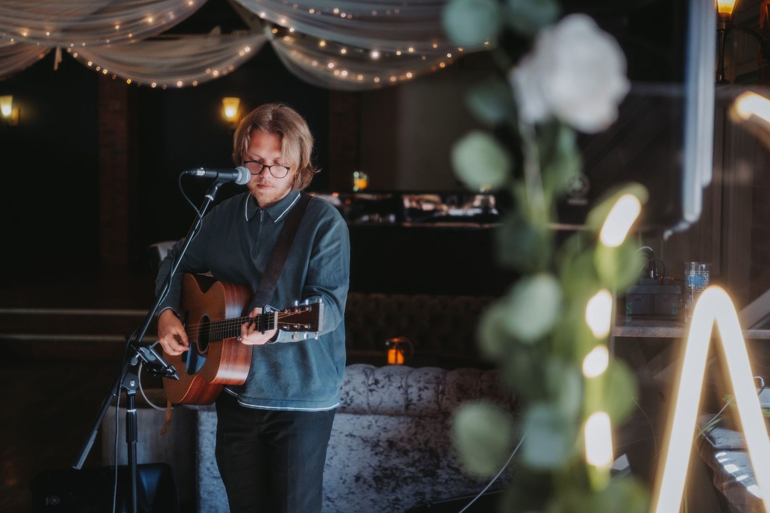 Musician performs with acoustic guitar on stage at wedding venue with string lights and moody lighting in background.