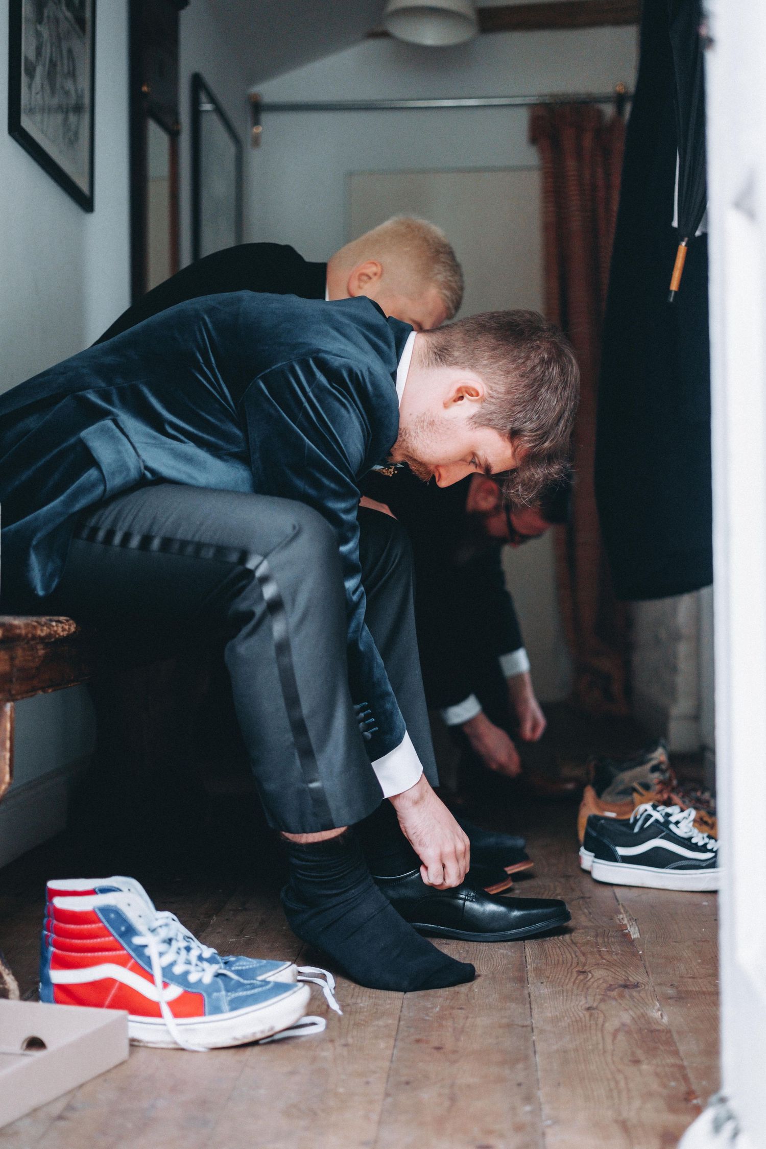 Someone puts on dress shoes while red and white Vans sneakers sit nearby on a wooden floor.