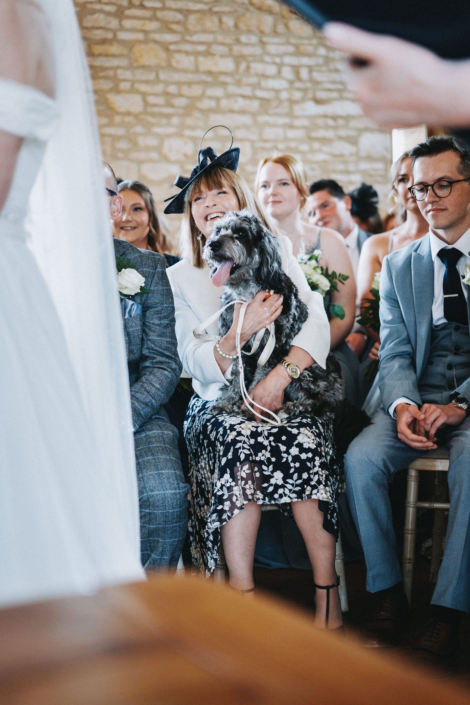 A wedding guest holds a small black and white dog during the ceremony in a stone-walled venue.