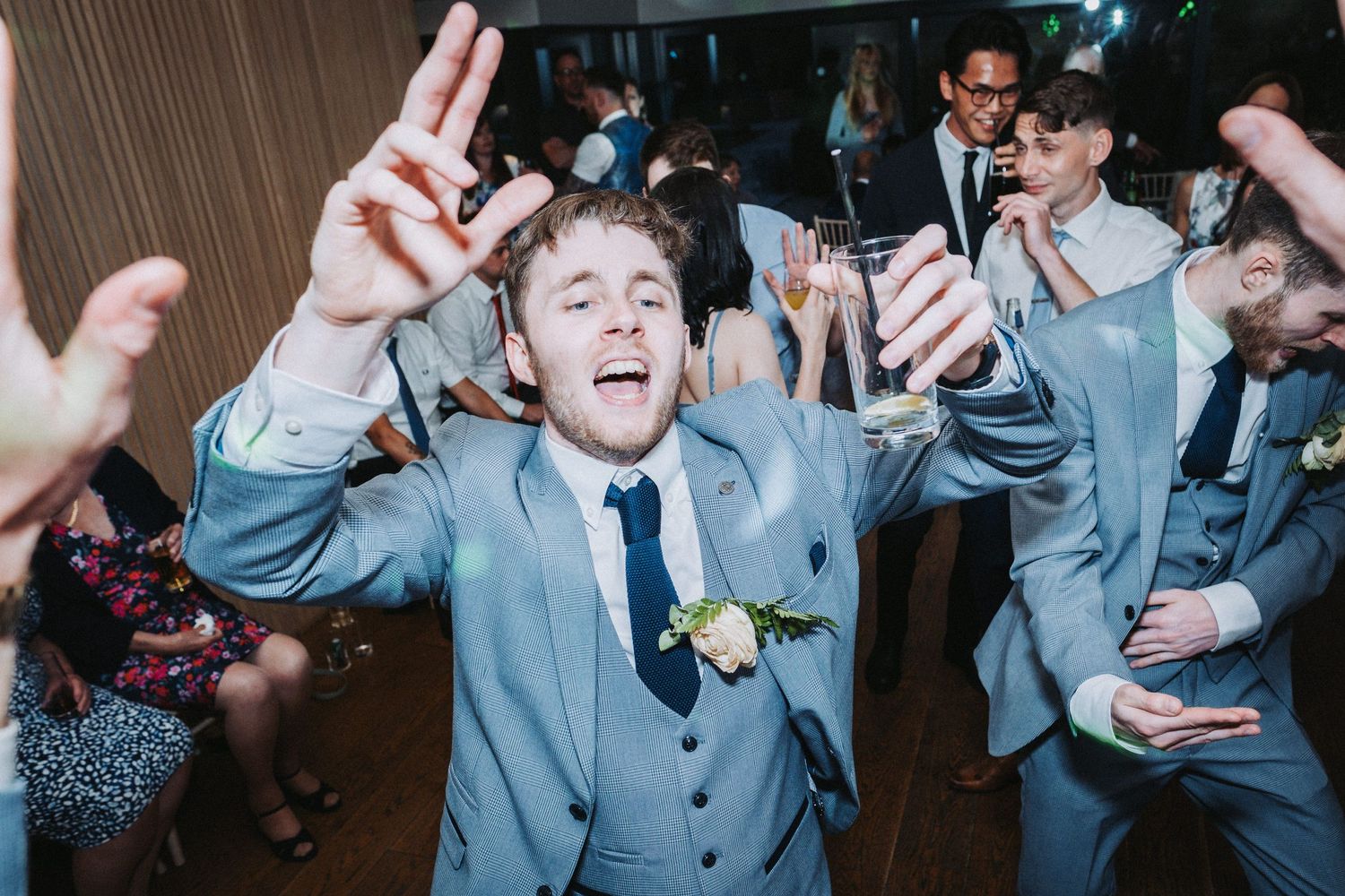 Wedding guest in light blue suit dances energetically with raised hands on crowded dance floor at reception celebration.
