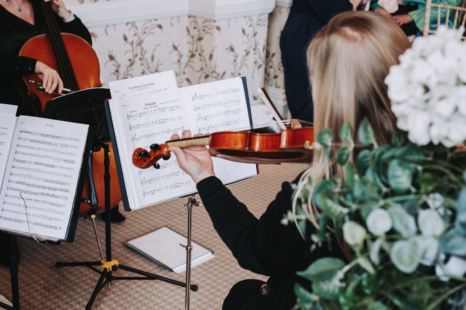 A musician plays violin next to sheet music and white floral arrangements.