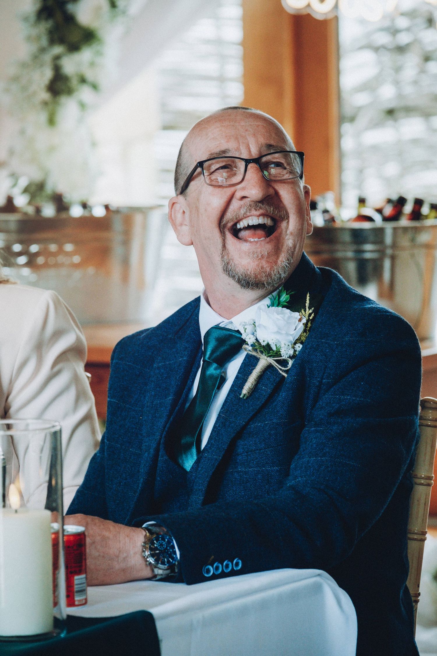 Wedding guest in navy suit and glasses laughs joyfully while wearing a white boutonniere.