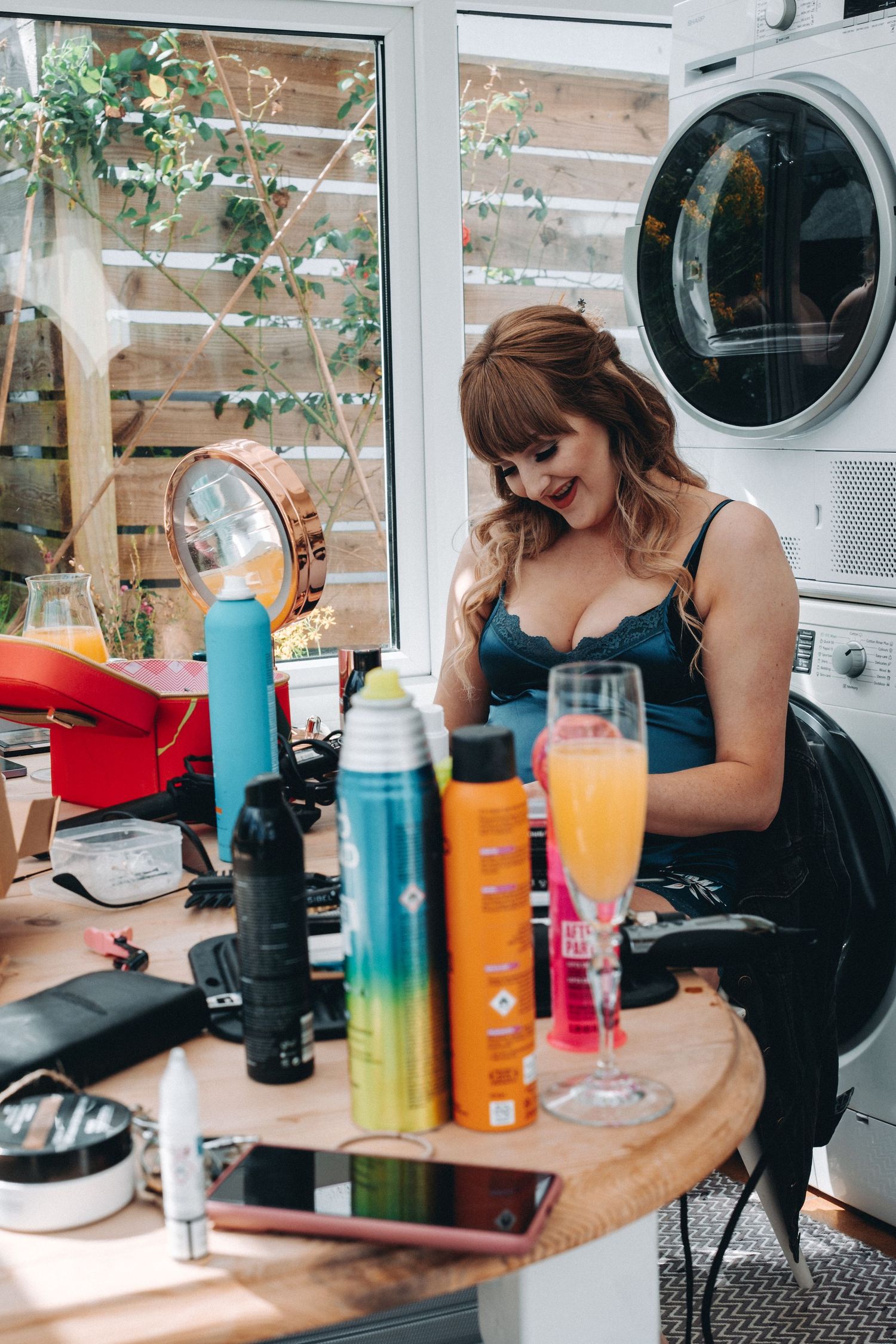 Bridesmaid with vanity setup with beauty products, colorful drink bottles and mimosa near a washer dryer unit with greenery visible outside.