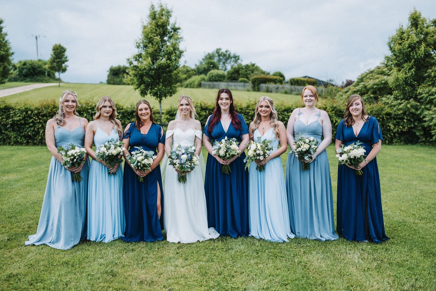 A wedding party in coordinated blue dresses stands together on a grassy hill holding floral bouquets.