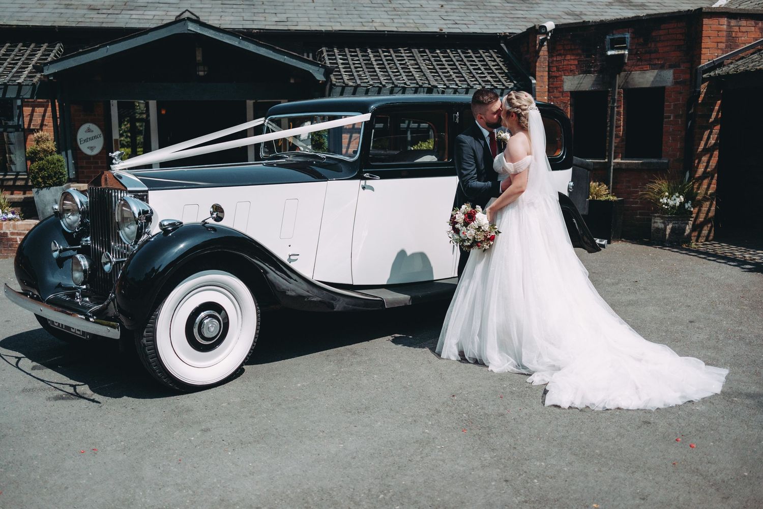 A classic white Rolls Royce vintage car with whitewall tires parked in front of rustic brick buildings.
