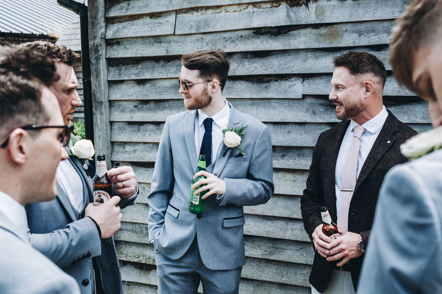 Wedding party in gray suits gathered outside a rustic barn sharing drinks and conversation.