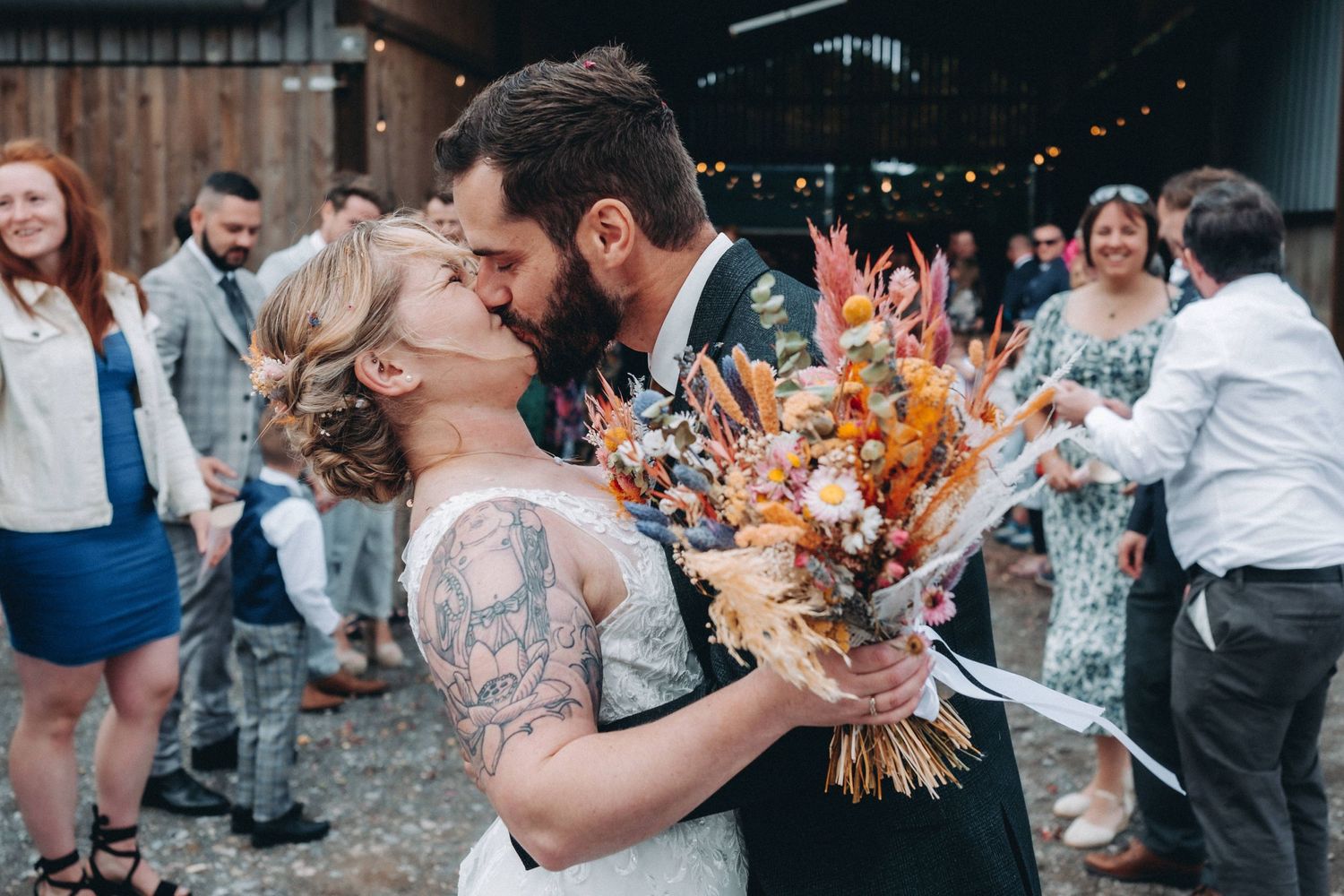 A couple shares their first kiss at a rustic barn wedding celebration while holding a vibrant orange dried flower bouquet.