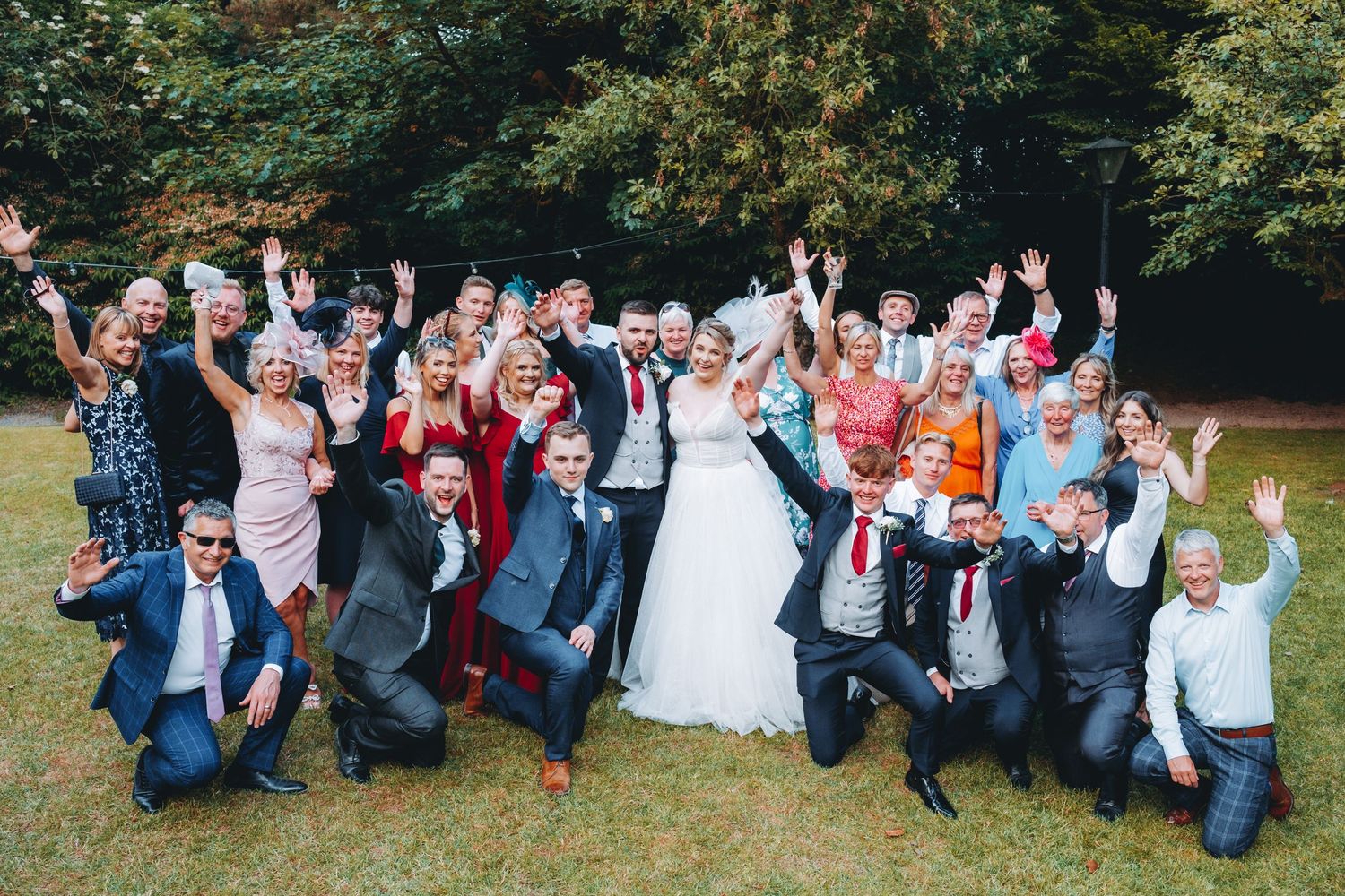 A large group of wedding guests pose together outdoors on a lawn with trees in the background.