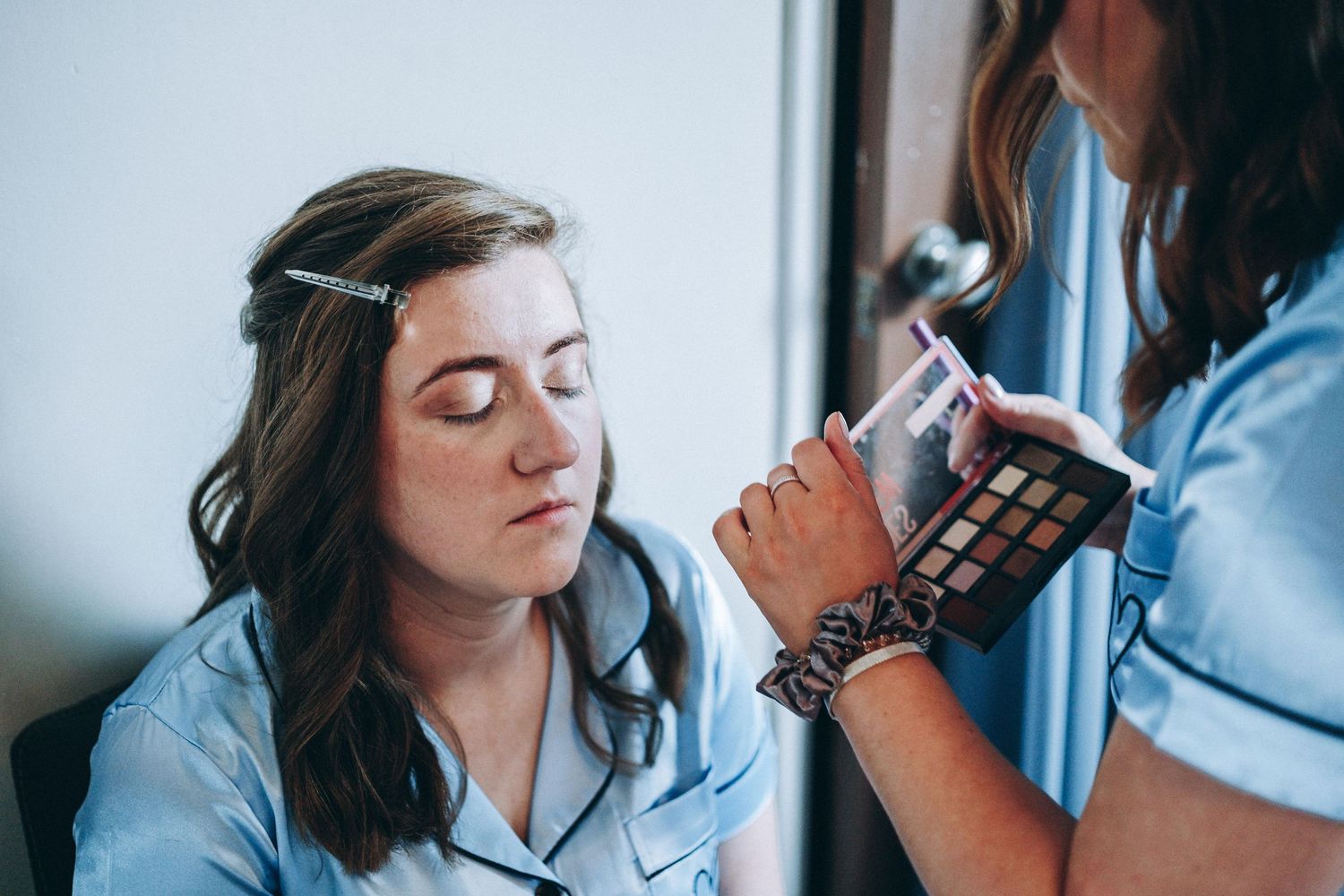 Makeup artist applying eyeshadow on a bridesmaid with a brush while sitting near a bright window.