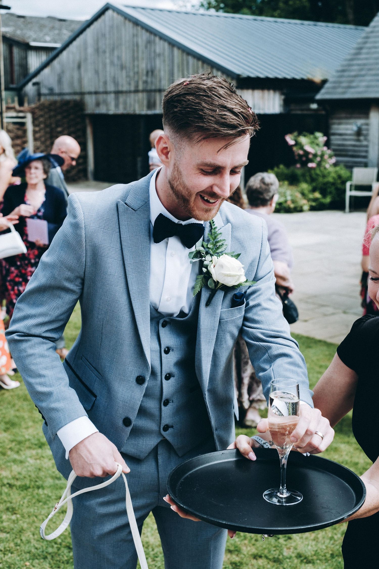 A person in a light blue suit carries champagne on a serving tray at an outdoor event near rustic buildings.