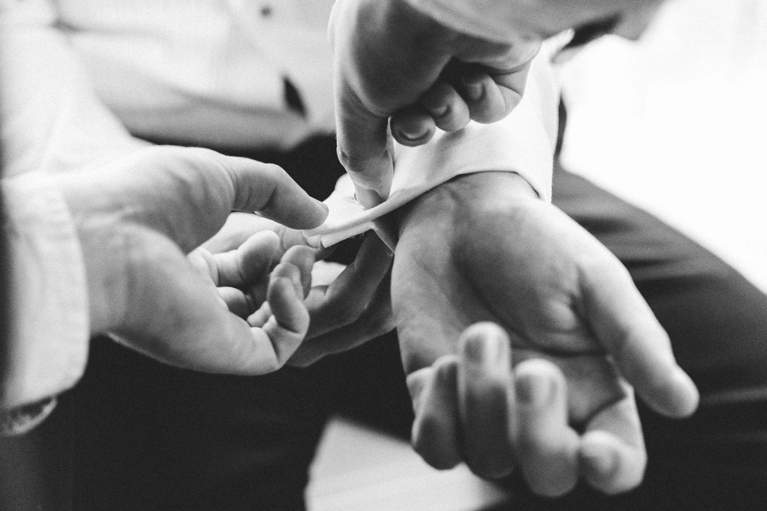Black and white image shows a groomsmen helping attach the grooms cufflinks.