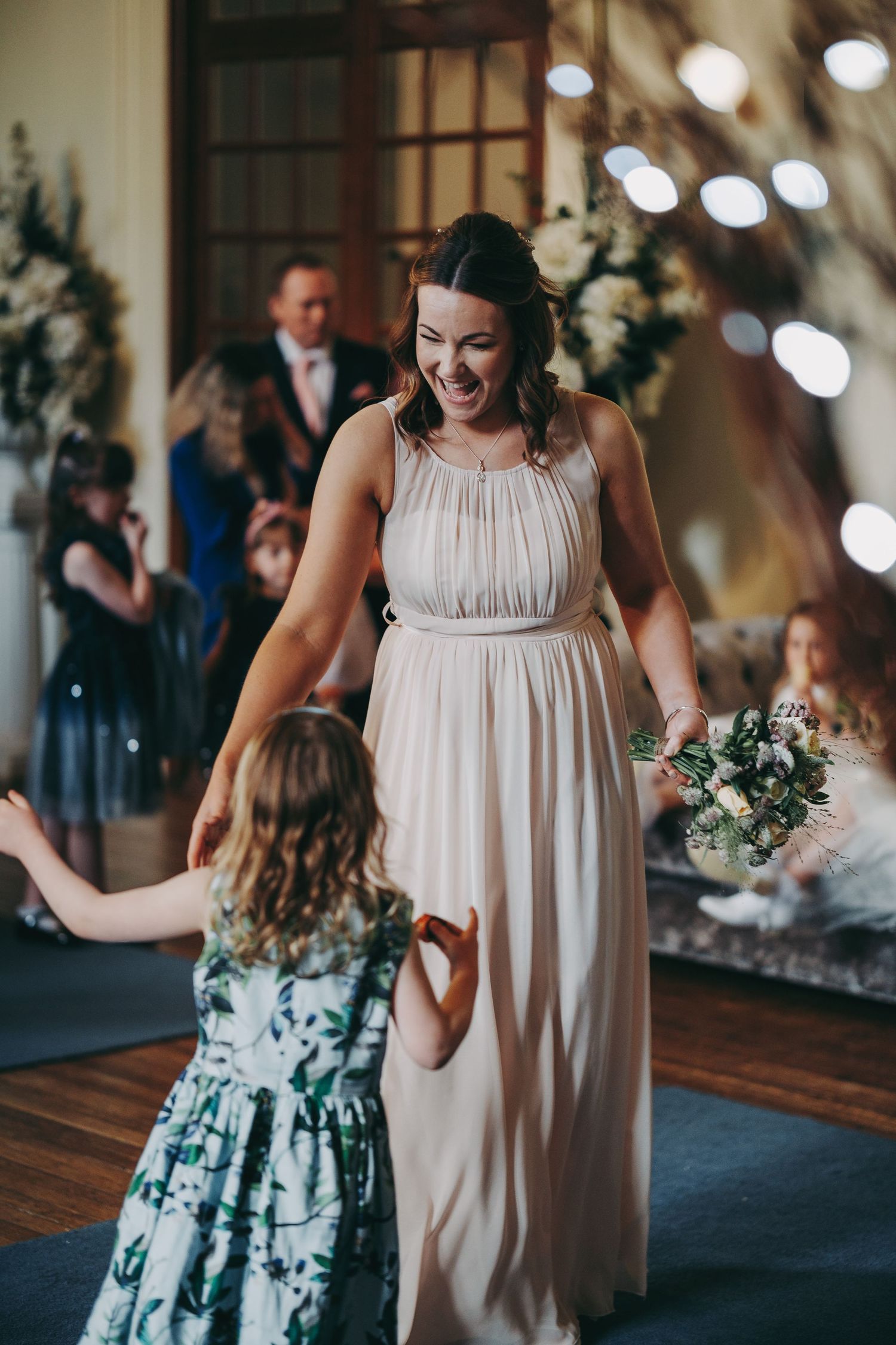 Bridesmaid in flowing blush dress shares a moment with young flower girl at reception.