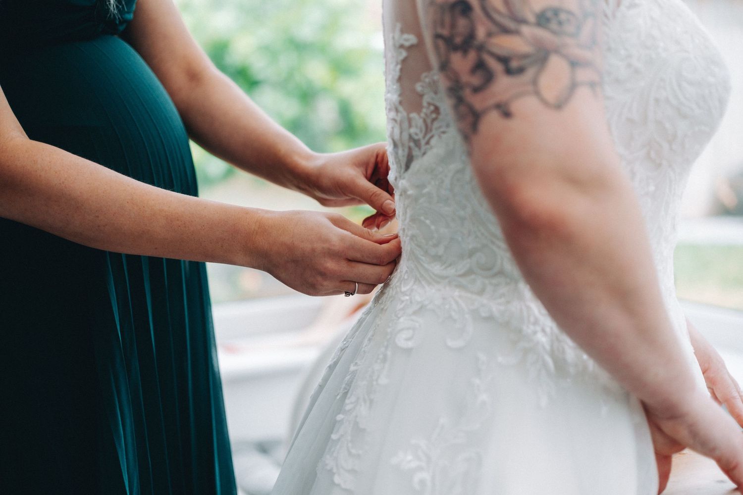 A person in a teal bridesmaid dress helps adjust details on a white lace wedding gown during preparation.