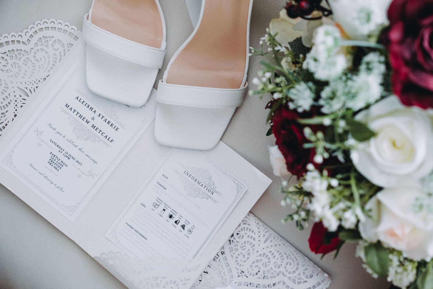 Wedding invitation cards with elegant white bridal shoes and red and white floral bouquet arrangement on light surface.