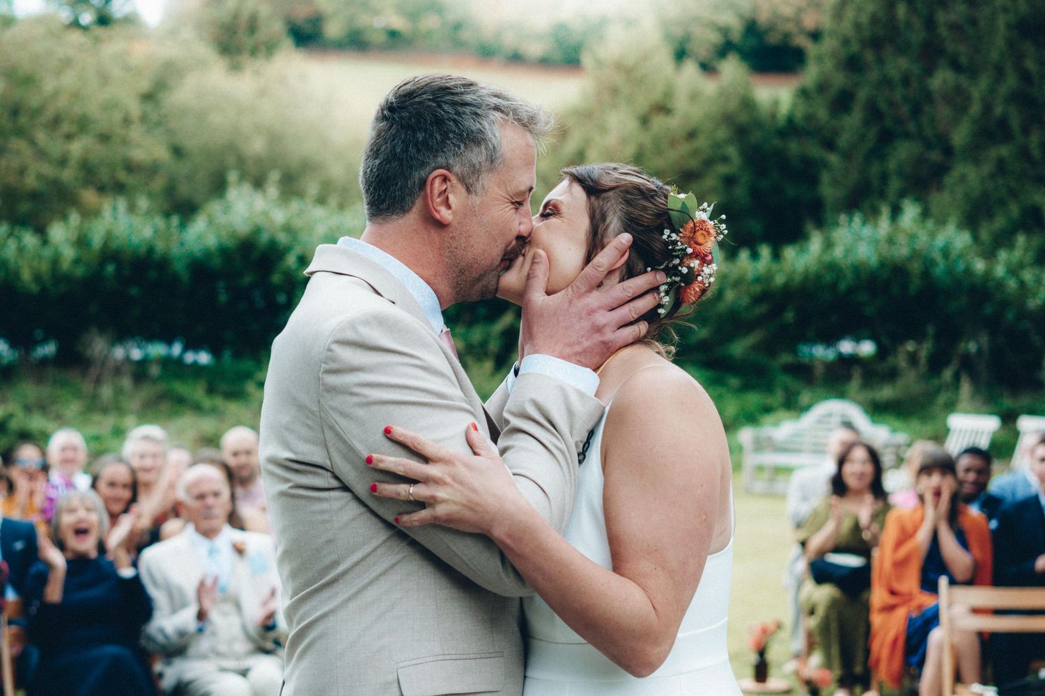 A couple shares a romantic kiss during their outdoor summer wedding celebration surrounded by guests and greenery.