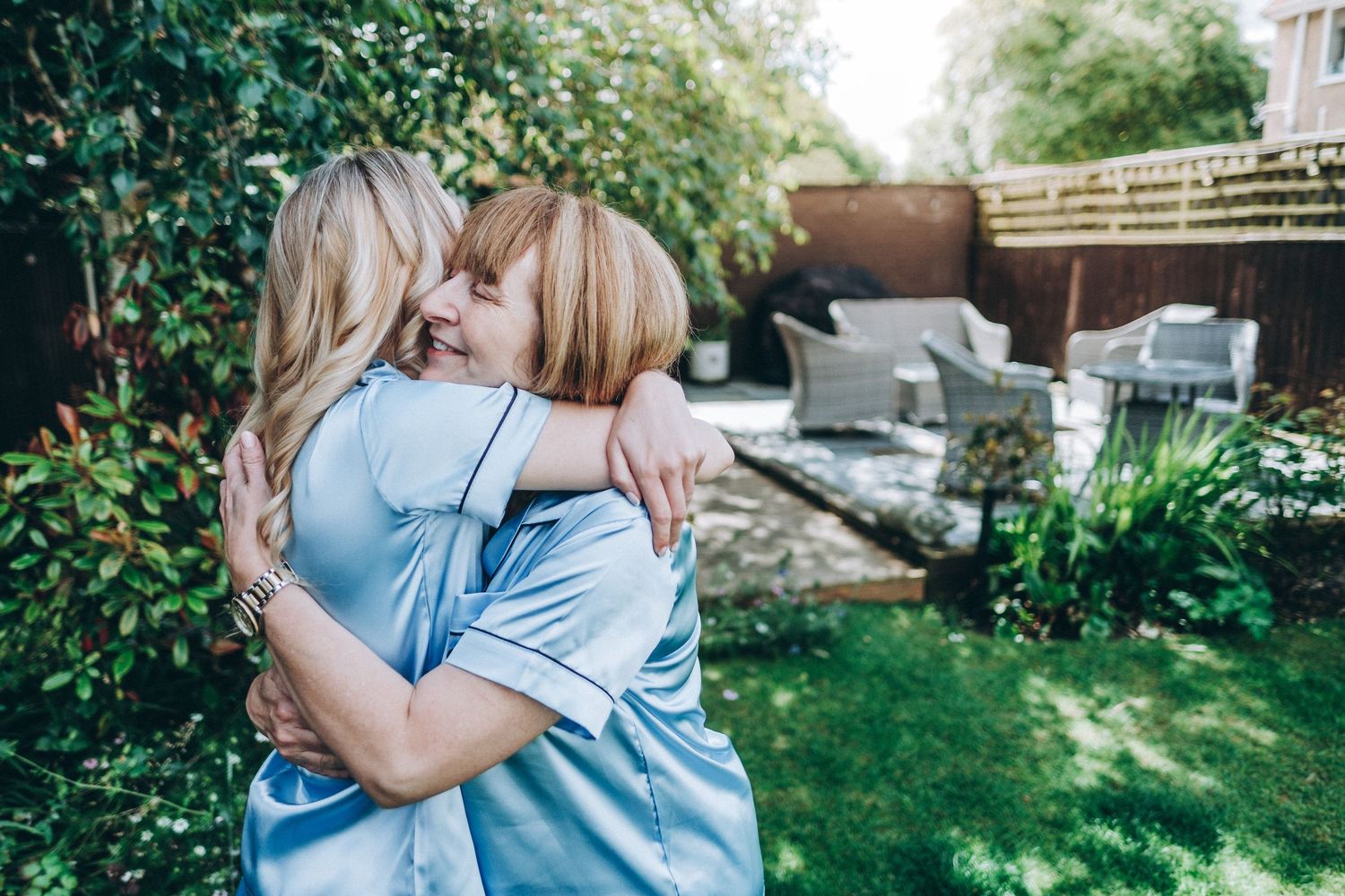 Two bridesmaids in matching blue robes embrace in sunny garden setting.