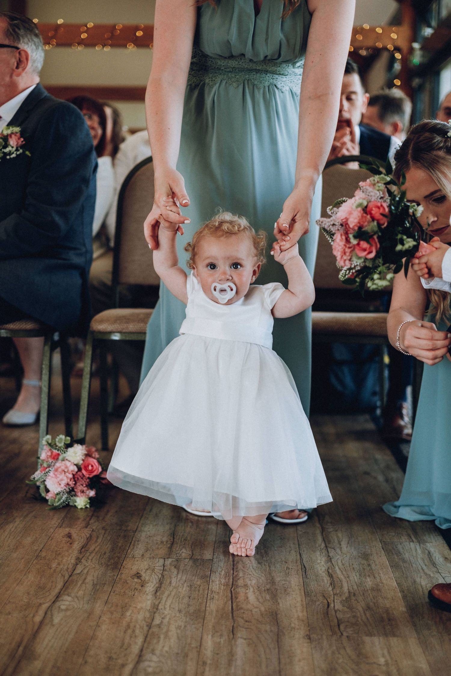 A small child in a white dress twirls on a wooden floor while bridesmaids in sage green dresses stand nearby.