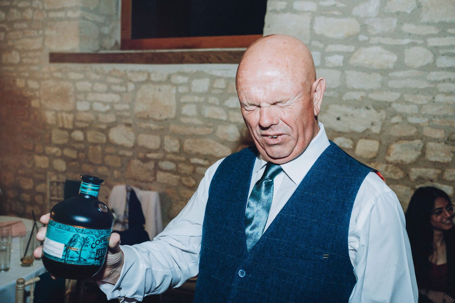 Guest in vest and tie enjoys a drink at stone-walled wedding venue.