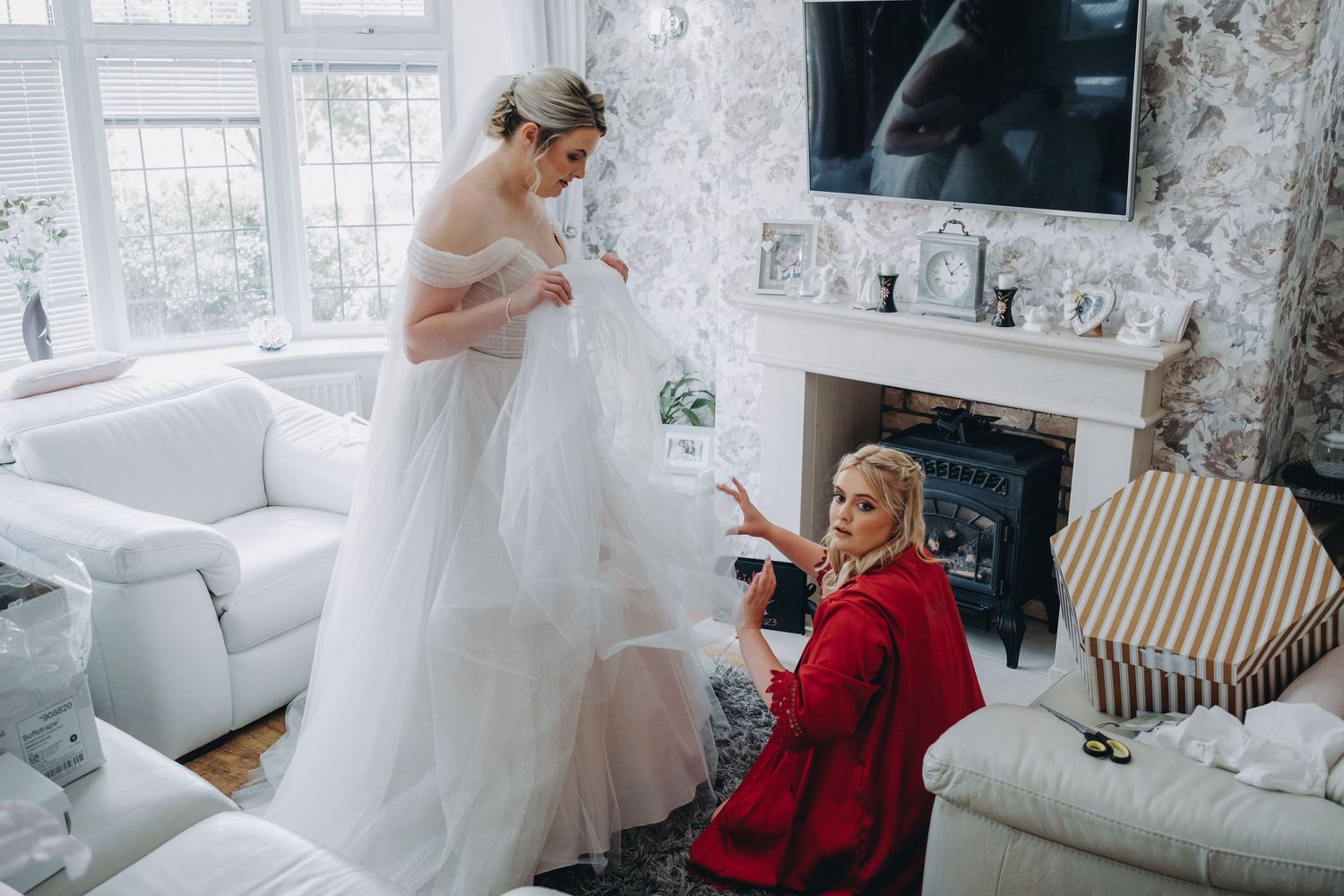 Bride and bridesmaid in a bright living room preparing for a wedding, wearing a white dress.
