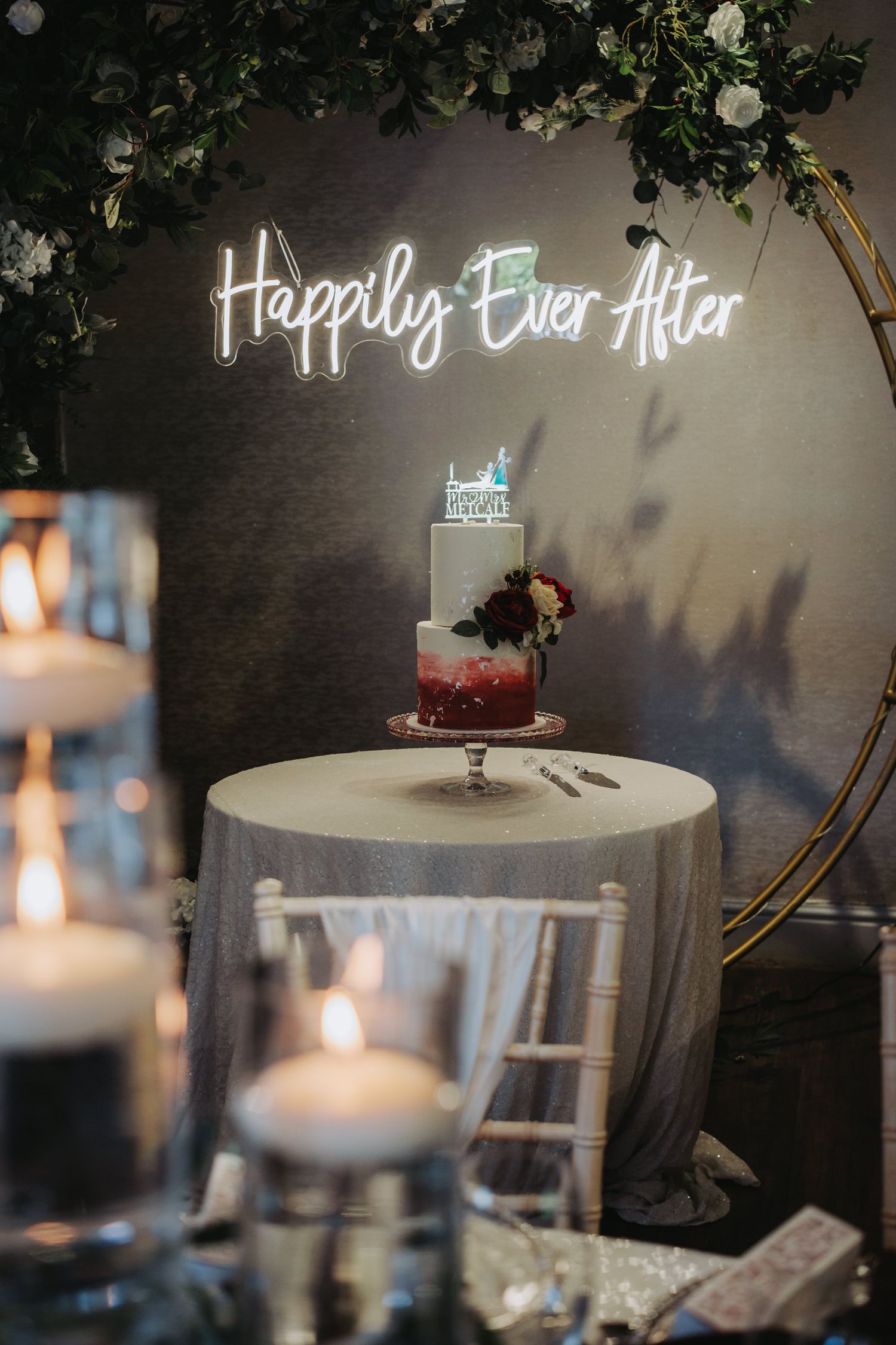 Wedding cake with neon 'Happily Ever After' sign glowing above it, decorated with candles and greenery in romantic setting.