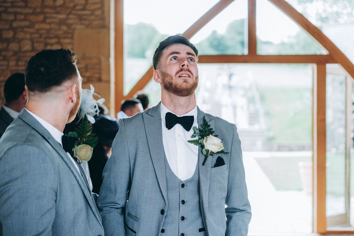A wedding party in formal attire gathers inside a venue with large windows and wooden beams.