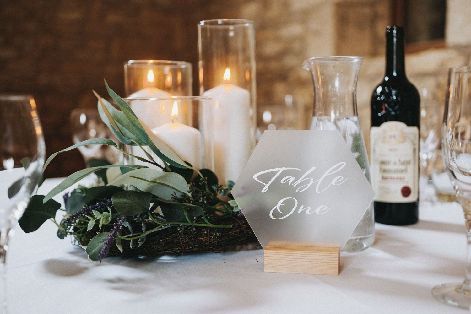 Elegant wedding table setting with white candles, greenery, and wine bottle against soft candlelight.