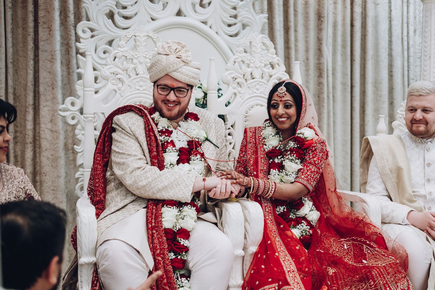 Couple in traditional Indian wedding attire seated on decorated stage during ceremony.