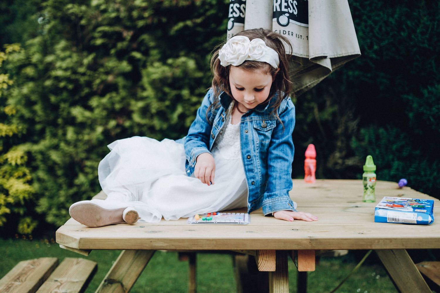 A white-dressed figure plays with toys on a wooden picnic table in an outdoor garden setting.