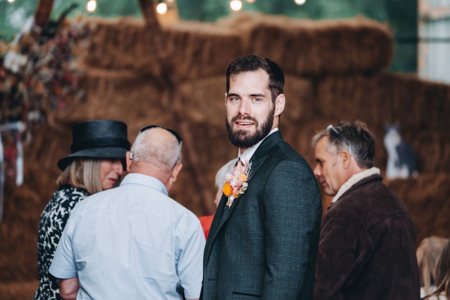 Wedding guests gather outdoors at a rustic venue as a groom in a dark suit with an orange boutonniere stands among them.