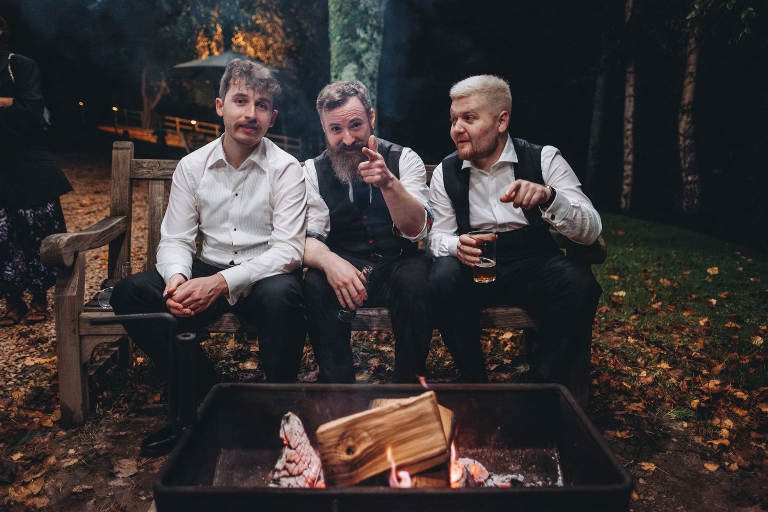 Three guests relax by outdoor fire pit during evening wedding celebration.