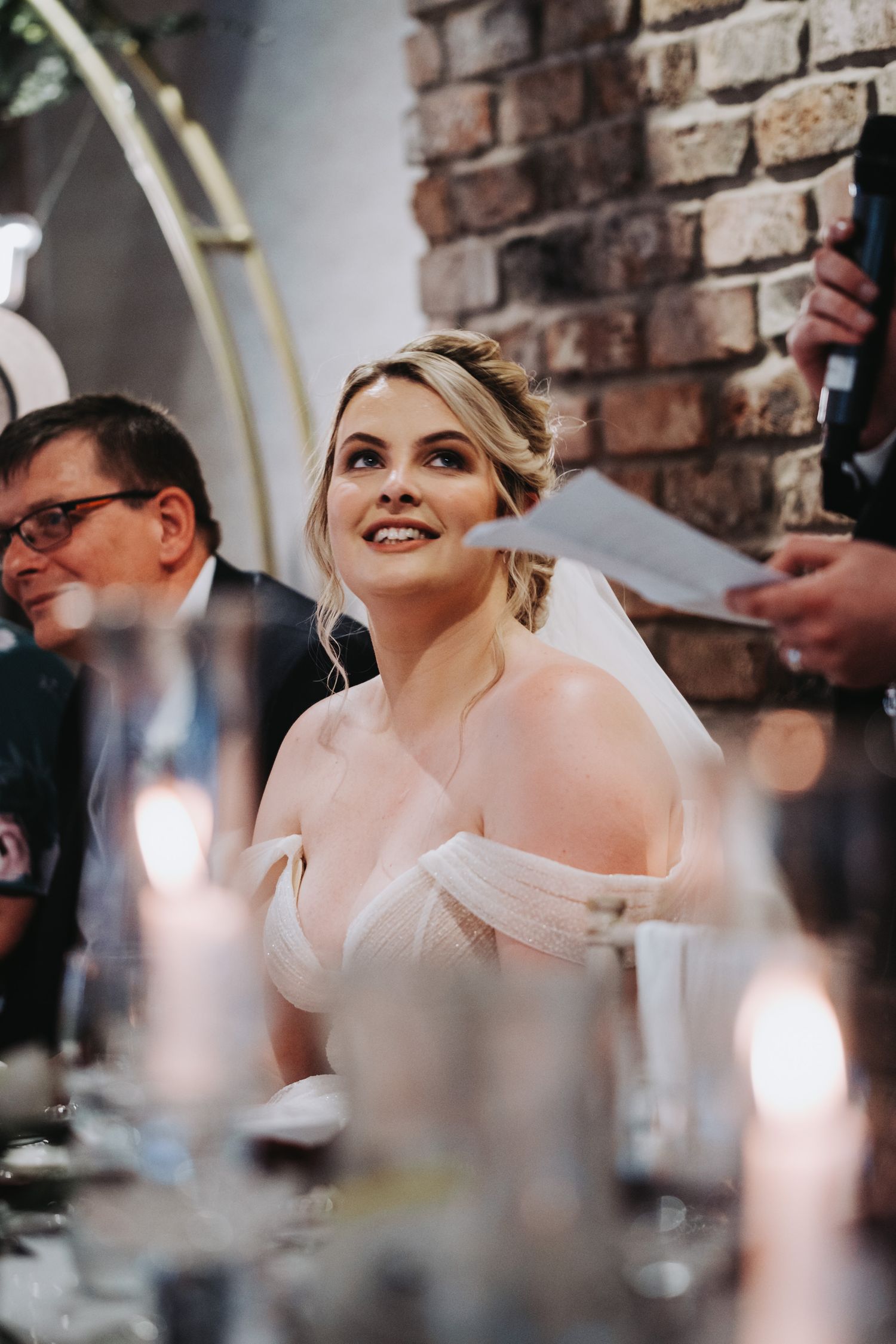 Bride in elegant off-shoulder wedding dress smiles during reception dinner at rustic brick venue with candlelight.