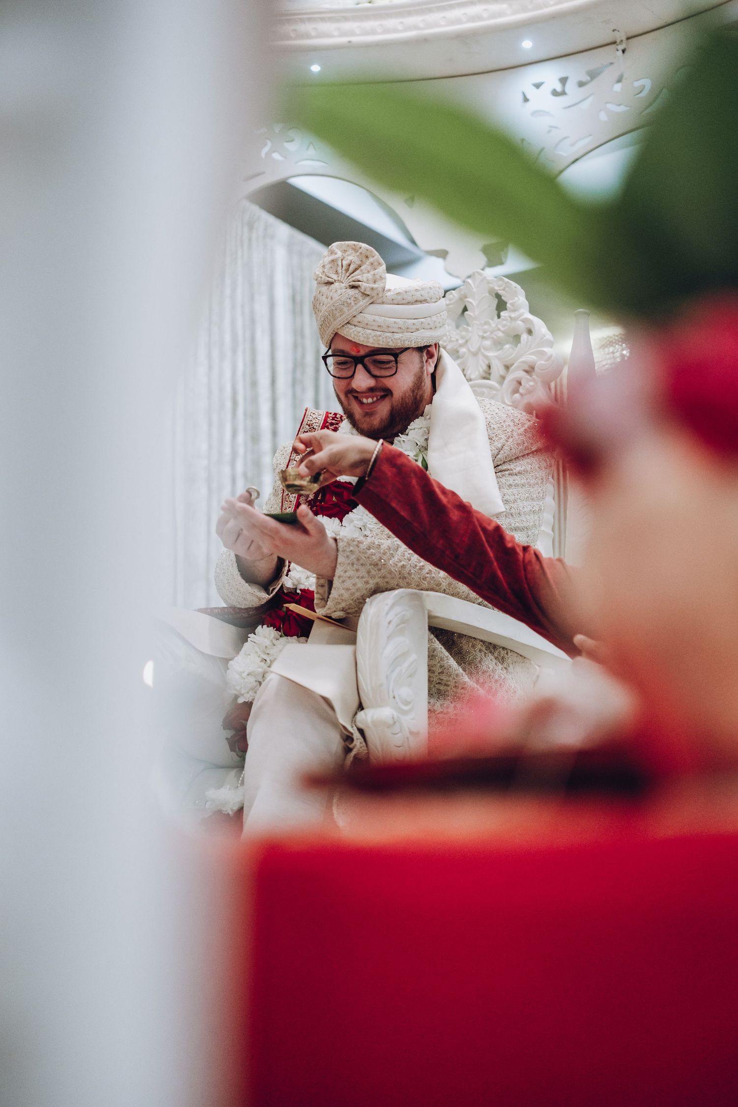 Wedding ceremony moment captured through foliage in an ornately decorated white interior setting.