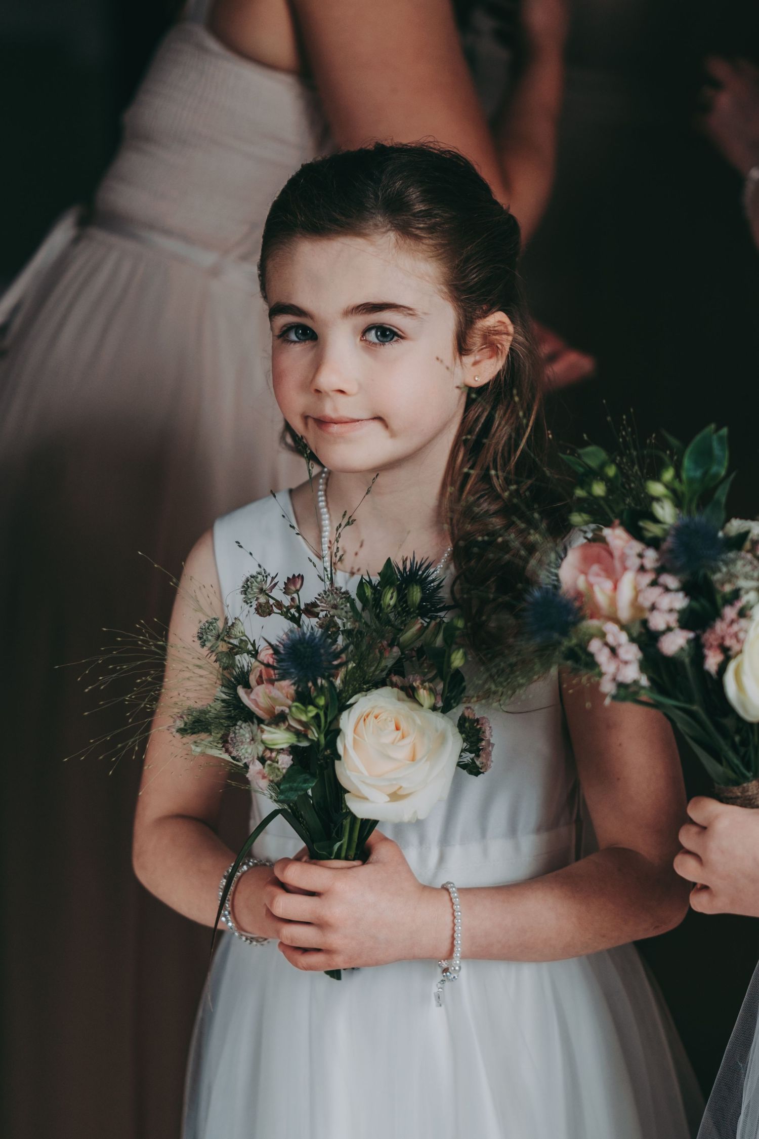 Young bridesmaid in a white dress holds a delicate bouquet of roses and greenery.