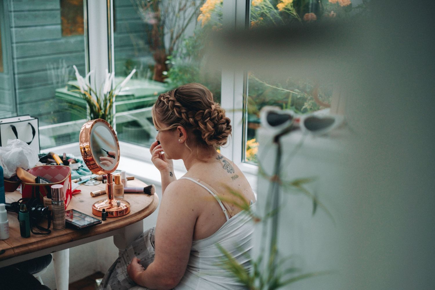 Bride in a white dress sits at a vanity table with makeup and beauty supplies while getting ready for a wedding.