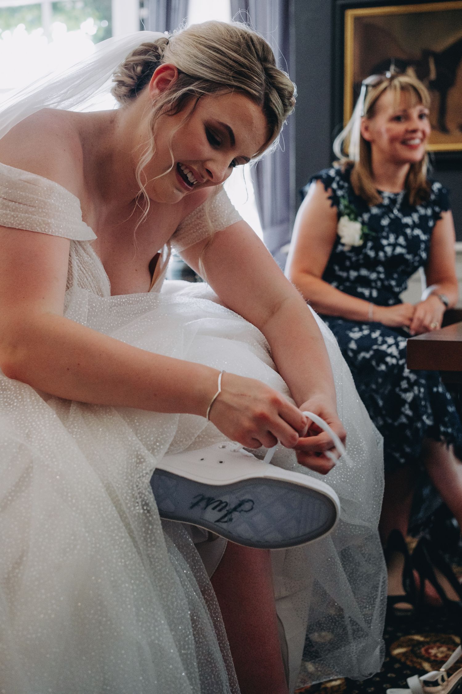 A bride in a white wedding dress with writing on the sole of her shoe while someone watches and smiles in the background.