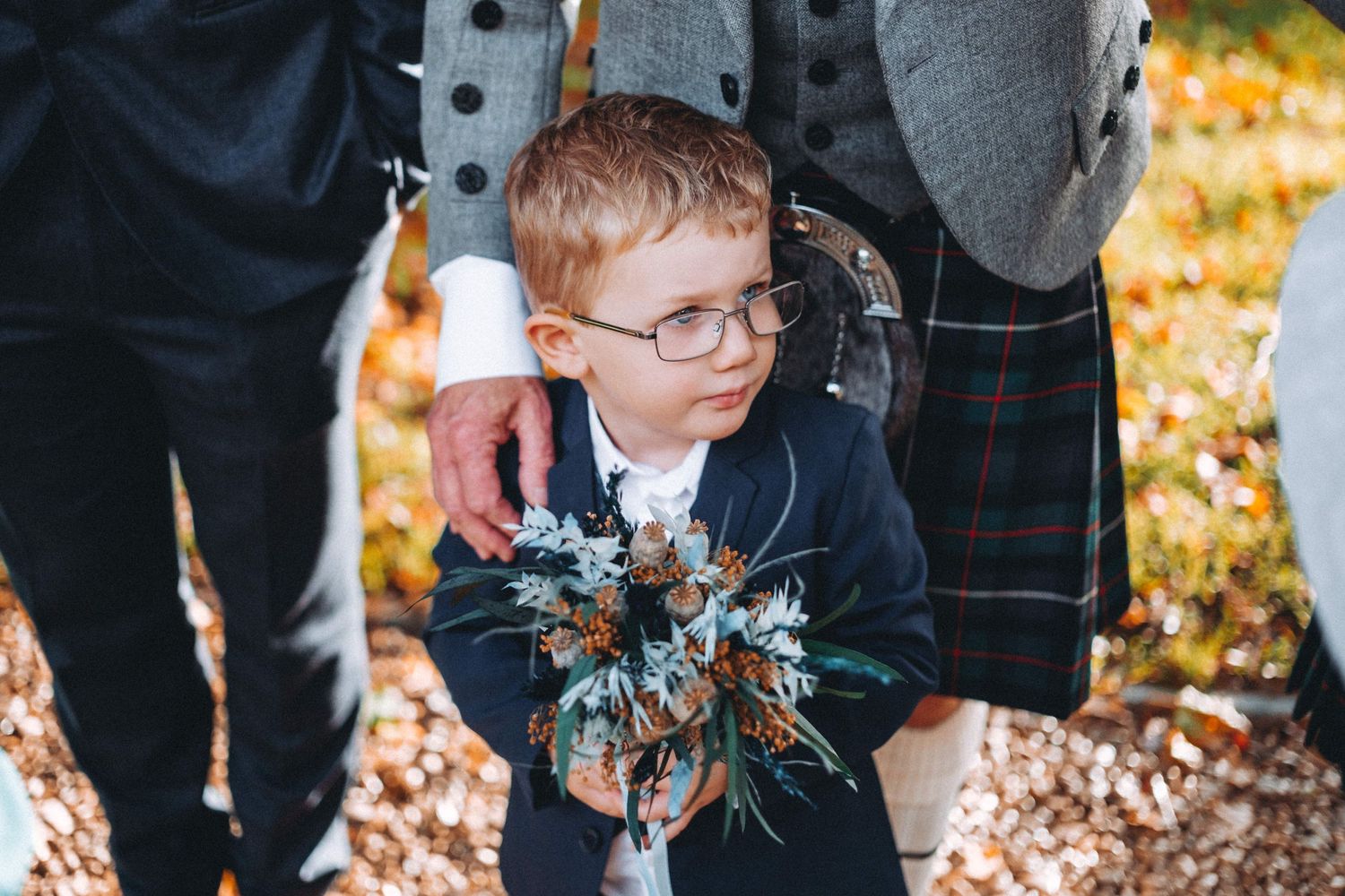 A young child in glasses and wedding attire holds a floral bouquet during an outdoor autumn ceremony.